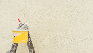 A paint roller sits in a bucket on top of a ladder in front on a painted exterior wall.