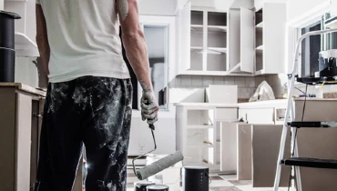 Photo of a paint-covered man holding a used roller brush looking at a kitchen being painted white.