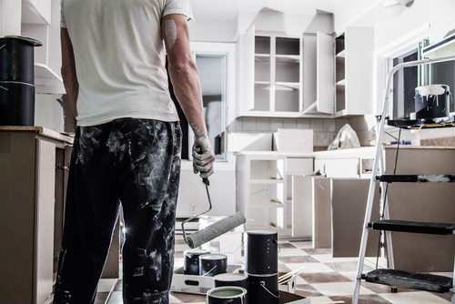 Photo of a man covered in paint and holding a used paint roller in a freshly painted white kitchen.