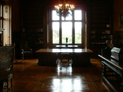 Elegant wood-paneled room with a large table, chairs, a chandelier, and a window overlooking greenery.