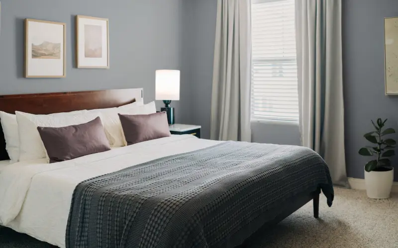 Modern bedroom painted a gray-blue with wood bed frame, minimalist decor, and window with curtains.